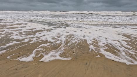 powerful ocean waves crash onto a sandy beach, creating foamy patterns under overcast skies, capturing the storm's intensity