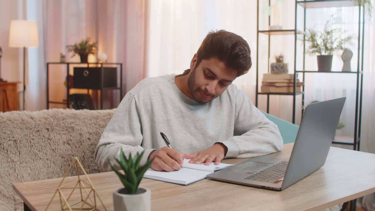 Young indian man student attending online class through laptop while taking notes at table at home