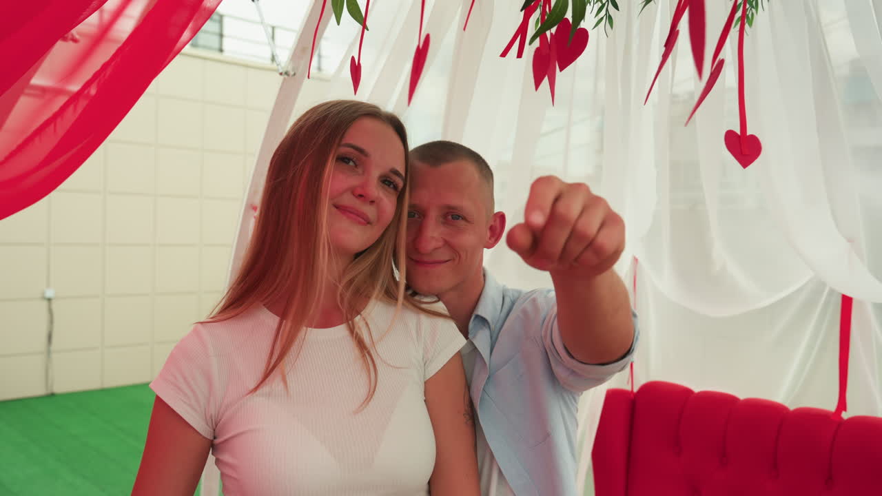 coppia caucasica in cabina di San Valentino, donna sorridente in abito bianco con capelli lunghi, uomo in camicia chiara in piedi vicino a decorazioni rosse a forma di cuore appese, baldacchino di drappeggio bianco trasparente, uomo su divano rosso imbottito