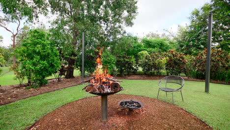 Jardín-Tropical-Rural-Con-Chimenea-Rugiente-En-Un-Claro-De-Tierra-Redondeado-Y-Sillas-De-Mimbre-Verde