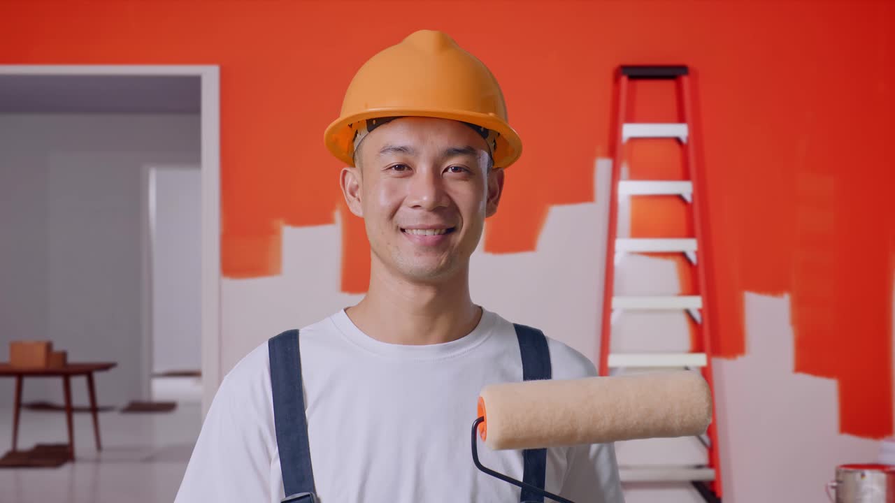 un pintor asiático con casco de seguridad de pie y sonriendo a la cámara con una pared de pintura en la habitación