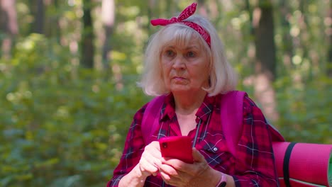 atractiva anciana abuela mujer mochilera planeando una ruta de viaje en el bosque en el teléfono móvil