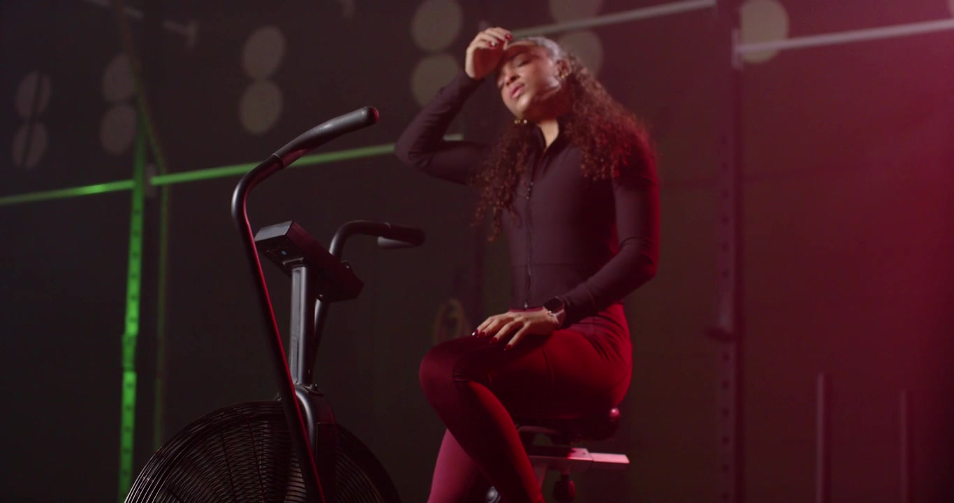 Donna afroamericana che pedala su una cyclette ad aria in una palestra poco illuminata, indossando una maglia con cerniera, allenandosi a intervalli.