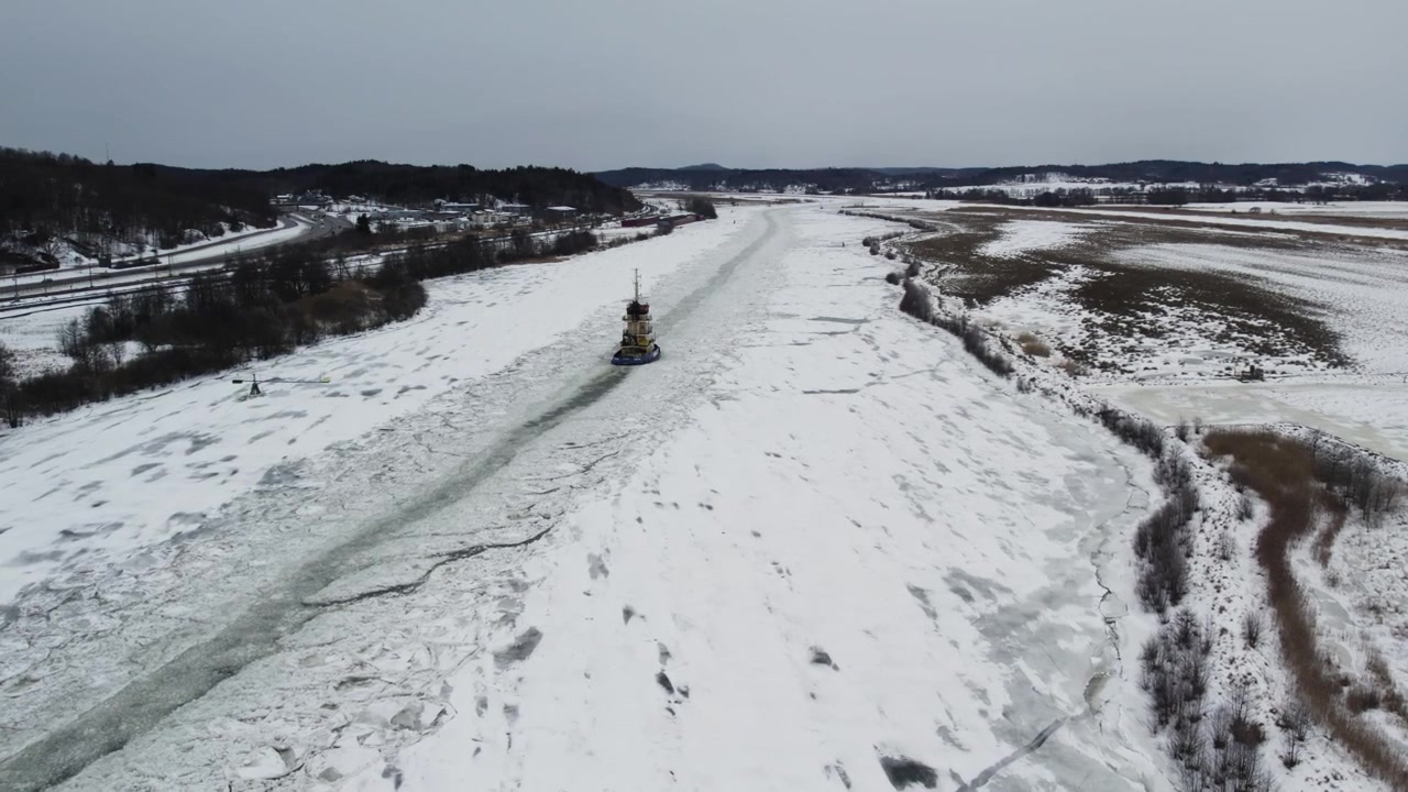 Ripresa aerea con drone che mostra una nave rompighiaccio che apre la strada nell'acqua ghiacciata attraverso il fiume Gotalv, circondato da campi innevati e paesaggio invernale in Svezia durante il giorno.