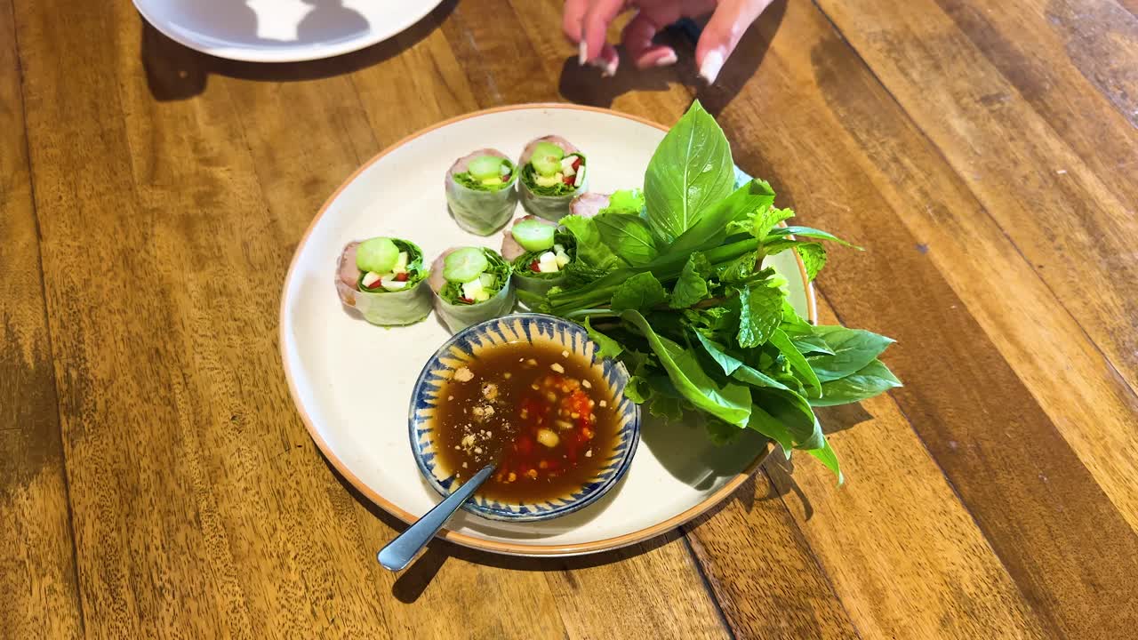 Hands assemble fresh Vietnamese spring rolls with herbs and dipping sauce on a wooden table in a well-lit restaurant