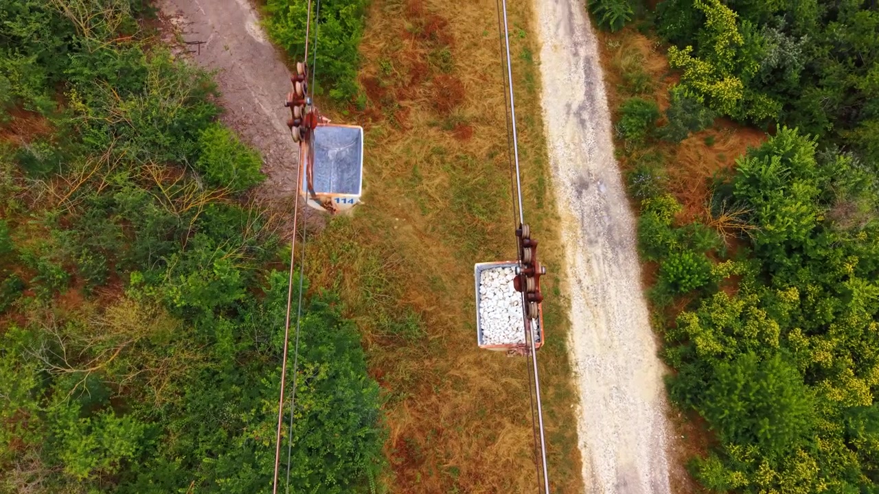 Industriel svævebane, der transporterer sten over en jordsti. Dronebillede ovenfra viser en rusten spand fyldt med hvide sten over grønne træer