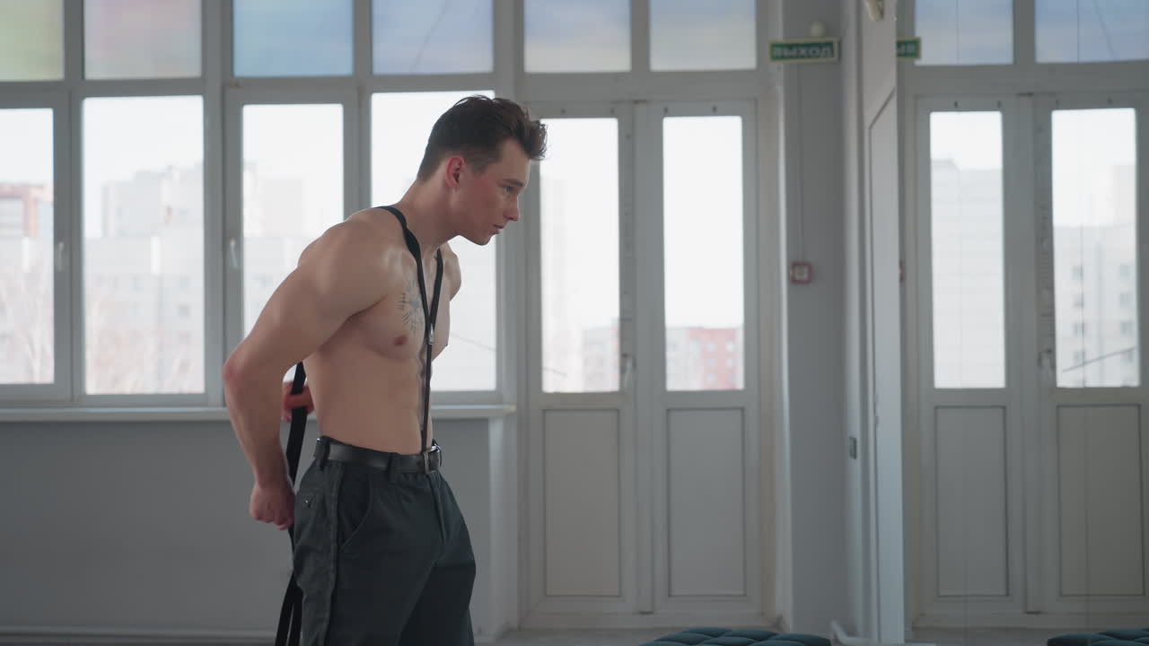 shirtless young man stands in front of mirror adjusting hook rope behind neck in bright indoor setting with large windows, focused on preparation for fitness routine under natural daylight