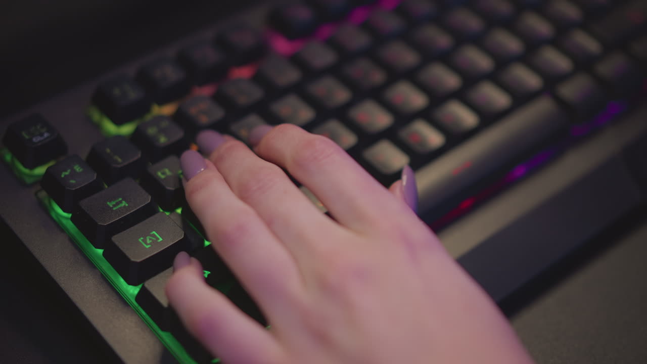 Close up hand view of woman with polished nails actively typing on rgb mechanical keyboard in moody ambient light setting, highlighting gamer or streamer environment with colorful tech detail