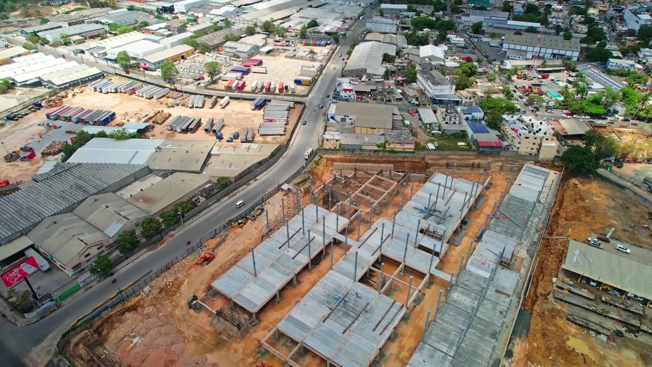 Ripresa aerea con drone dall'alto che vola in avanti e si inclina verso il basso sopra un grande cantiere edile per un edificio commerciale, mostrando pilastri in cemento, travi in acciaio e dintorni industriali