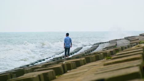 Hombre-Solitario-Caminando-A-Lo-Largo-De-Una-Defensa-Marítima-Mientras-Las-Olas-Rompen,-Concepto-De-Soledad