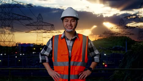 ingeniero masculino asiático con casco de seguridad sonriendo a la cámara mientras está de pie con los brazos akimbo cerca de la torre de alto voltaje