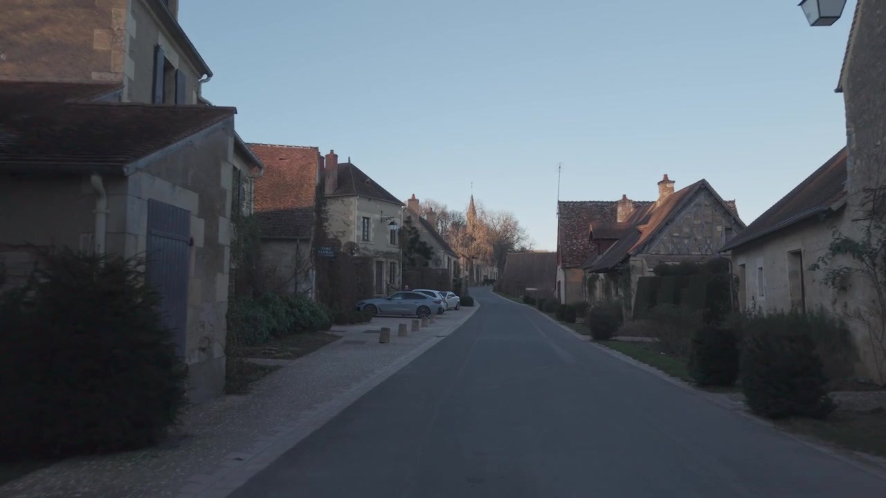 An aerial view looks down a narrow stone village street lined with parked cars in the Loire Valley, France.
