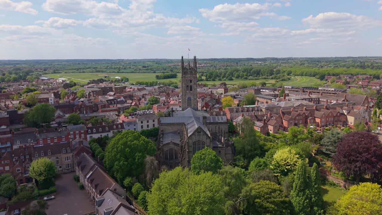 Dron se aproxima a la Iglesia de St Mary en Warwick en un día soleado, revelando su arquitectura histórica y el pueblo circundante bajo la luz clara del día.