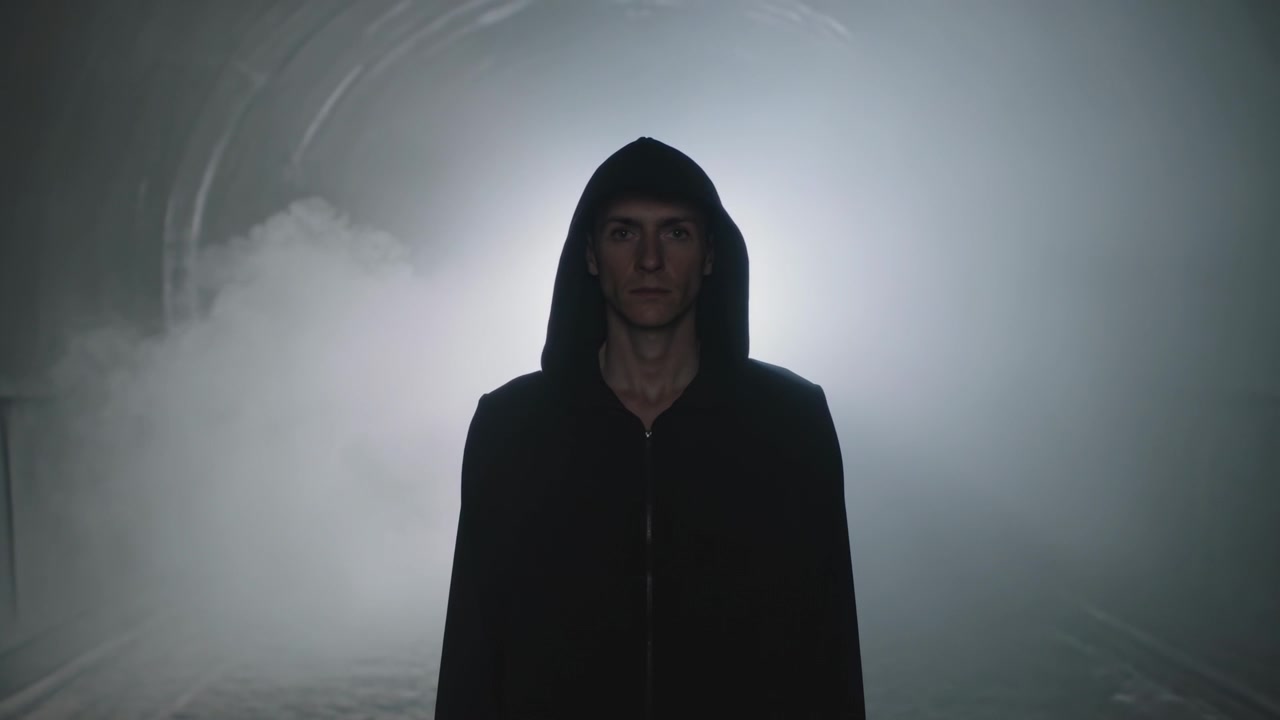 Hombre de pie con sudadera negra frente a la lente en un túnel arqueado, contraluz ascendente, niebla formando un halo alrededor de la figura.