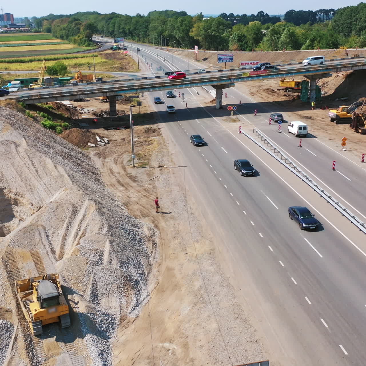 Trucks are working near the highway. Cars moving on the road. New bridge over the road with trucks and cars moving on it. Machinery for making new road in summer. Aerial view.