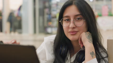 elegant lady with long dark hair and floral hand tattoo gently adjusts glasses with soft smile, resting head on hand while seated indoors, with blurred colorful background