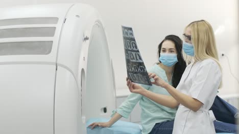 woman doctor radiologist explains the results of ct scanning for young female patient, showing the snapshot with images, observing and analyzing ct scan in modern clinic beside modern ct scanner