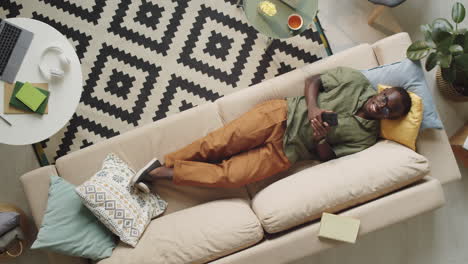 Cheerful-Black-Man-Lying-on-Sofa-and-Using-Smartphone