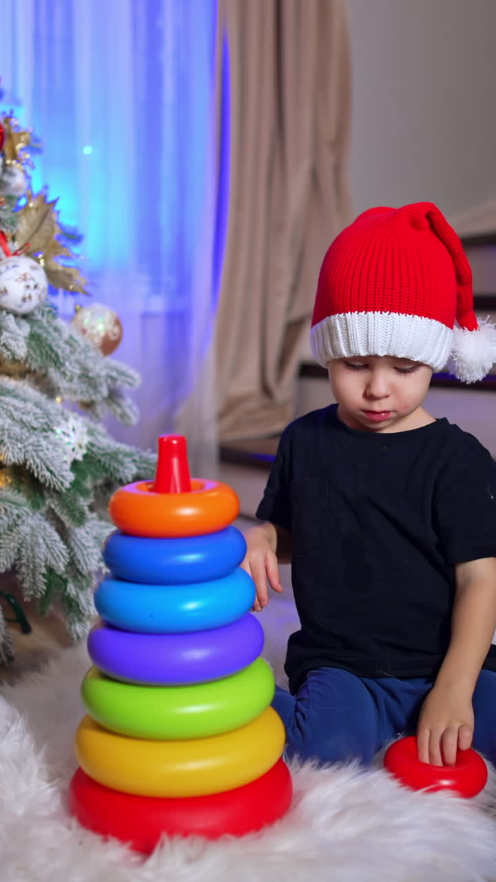 Lovely baby assembles a pyramid and then pushes it fall. Toddler in Santa cap playing near the Christmas tree. Vertical video