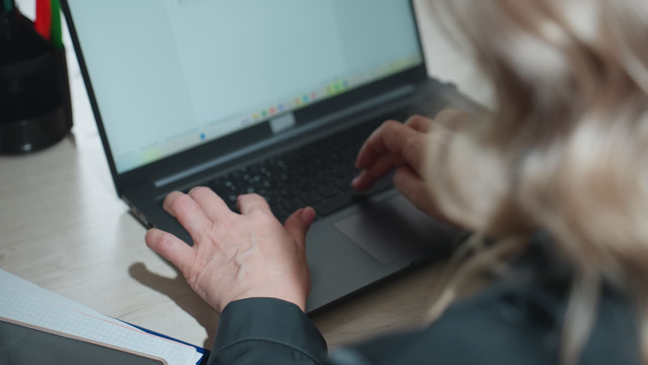 Rear view of woman typing on laptop in modern office setting with sleeves rolled and focused posture, showing hands on keyboard and blurred view of screen