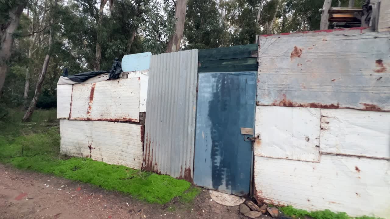 Ripresa in movimento che passa accanto a una parete di metallo ondulato rattoppata con ruggine e una porta blu, circondata da alberi e un sentiero sterrato a Barbate, Cadice.