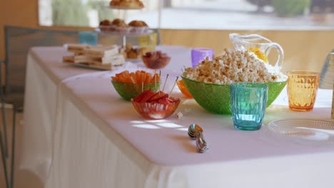 mesa con dulces para una fiesta en verano, con una bandeja con croissants, sándwich, palomitas de maíz, crudités, vasos de colores y mantel blanco moviéndose por el viento