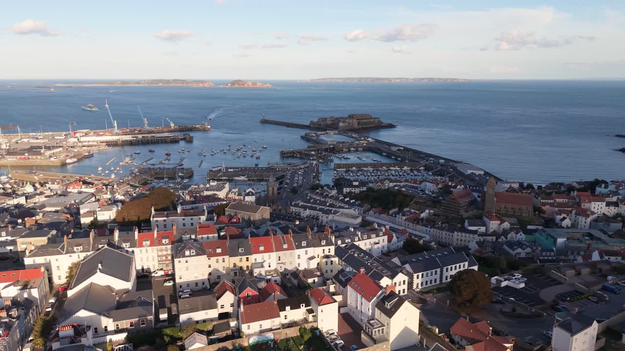 High circling flight over rooftops of St Peter Port Guernsey centring on harbour and Castle Cornet with Herm Sark and Jethou in the distance in bright afternoon sunshine on calm day