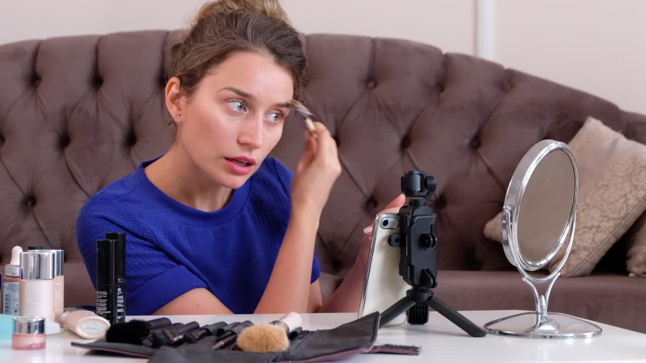 Woman in a blue T-shirt filming herself while doing a make-up tutorial at home