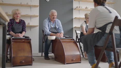 medium shot of middle aged ceramic artist teaching group elderly caucasian woman and senior man how to wedge clay sitting at desk in art studio. people enjoying talking at work