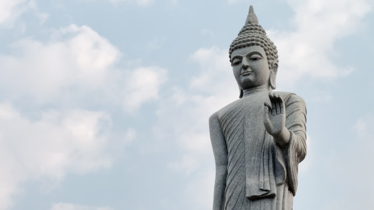 タイの公立寺院にある石仏像を、青空を背景に下から撮影したローアングルからの眺め。東南アジアにおける仏教、精神性、平和、瞑想の象徴。
