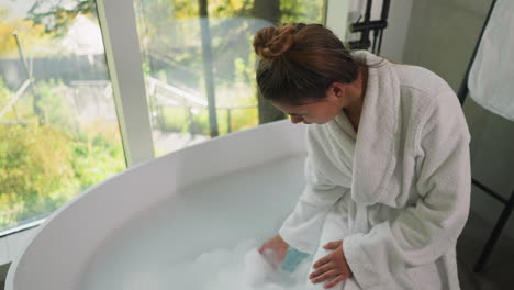 dama toca espuma sentada en el borde de la bañera. mujer joven en bata de baño suave juega con baño de burbujas esperando que el agua se enfríe. procedimientos de spa en casa