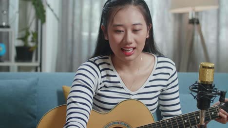 close up of asian woman reading comments on laptop and speaking to camera while live stream playing a guitar at home