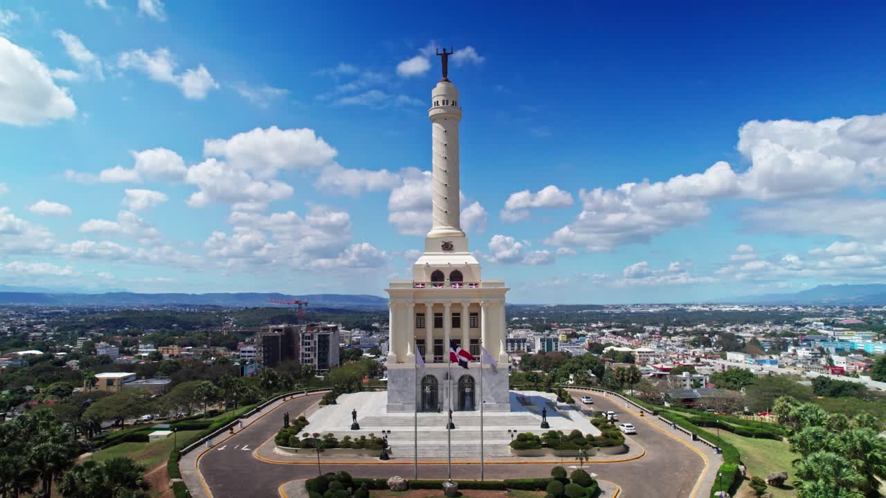 Ripresa a piedistallo con drone che inizia dalla statua equestre, rivelando il Monumento agli Eroi della Restaurazione. Ampia vista aerea dello skyline della città di Santiago sotto un cielo azzurro e soleggiato.