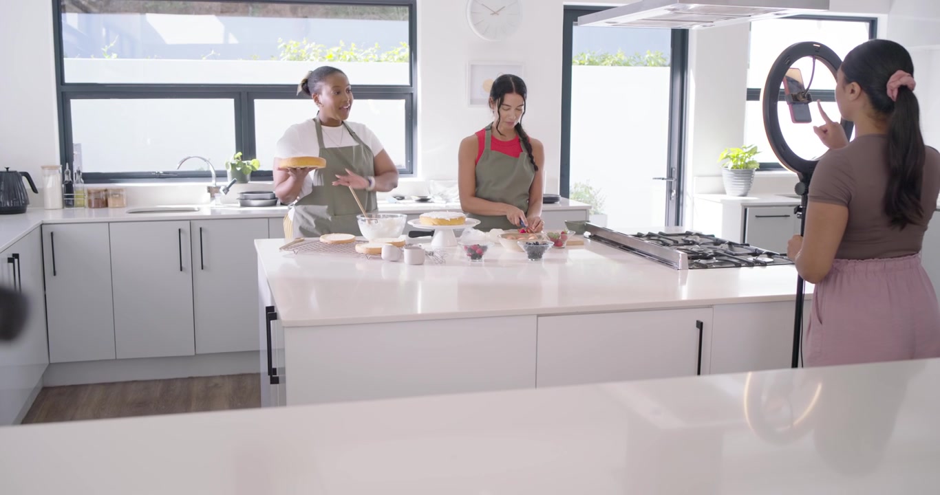 Diverse conduttrici con la telecamera in funzione, che salutano e sistemano una torta sull'isola della cucina per la registrazione.