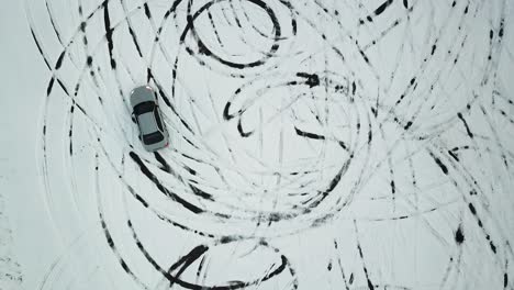 top down aerial of an older silver sedan doing power drifts on a snowy parking lot