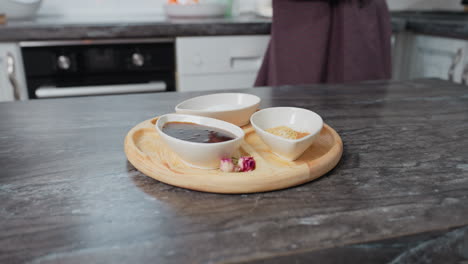 partial view of woman in elegant gown setting up lunch on black sleek table, stylish ceramic bowls with sauces and spices placed on wooden tray with decorative dried flowers