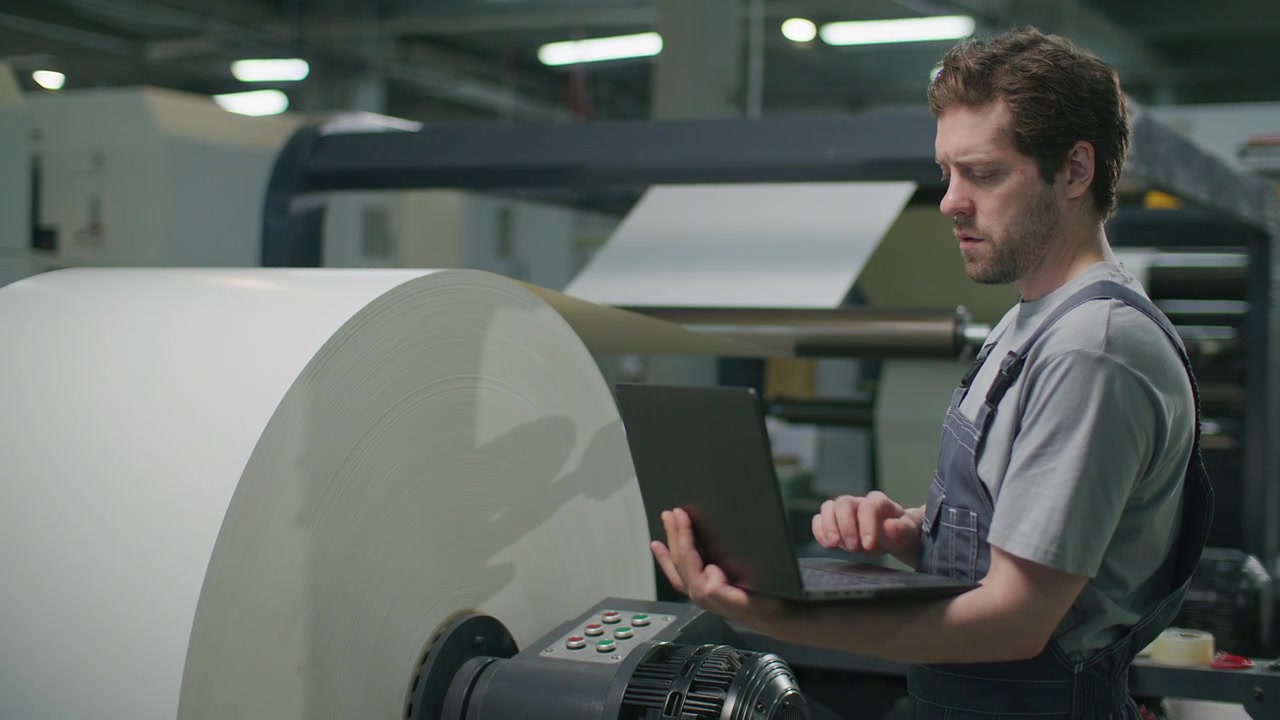 Industrial Operator Using Laptop to Monitor Web-Fed Press in Printing Factory