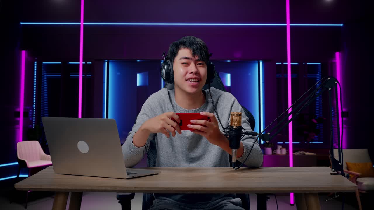 un hombre asiático sonriente jugador con auriculares y computadora agitando la mano y señalando el dedo índice luego jugando un juego de teléfono móvil en la habitación iluminada por luces de neón en retro