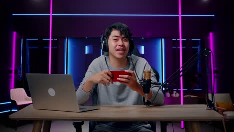 um jogador de videogame asiático sorridente com fones de ouvido e computador agitando a mão e apontando o dedo indicador, em seguida, jogando um jogo de telefone móvel na sala iluminada por luzes de néon em retro