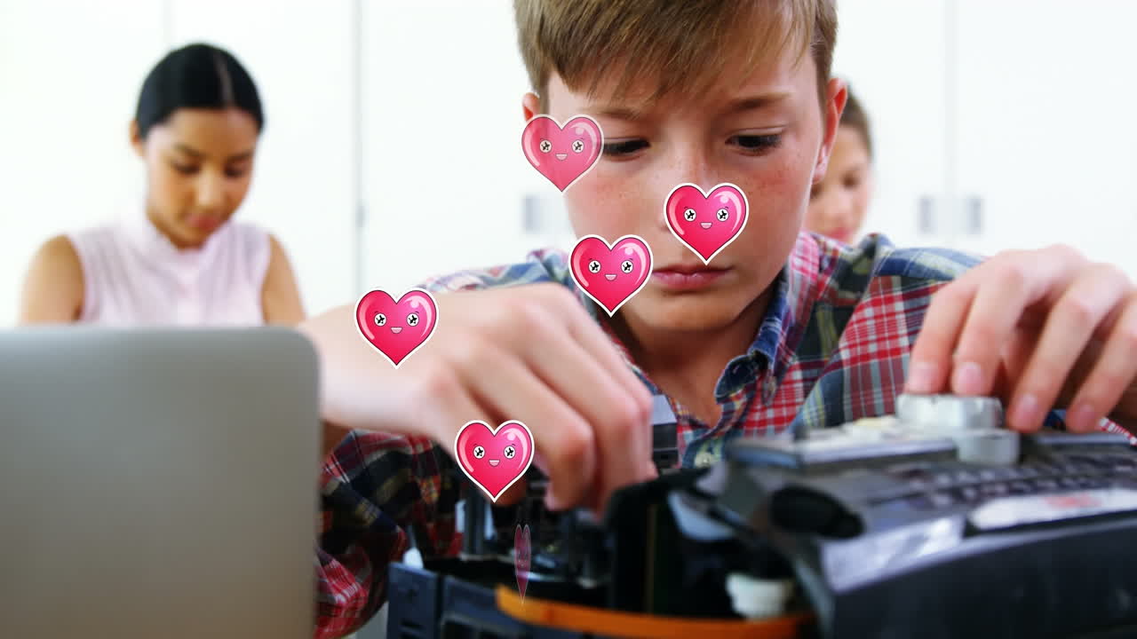 학교에서 교실에서 컴퓨터를 수리하는 백인 소년 위에 떠다니는 여러 개의 분홍색 심장 아이콘