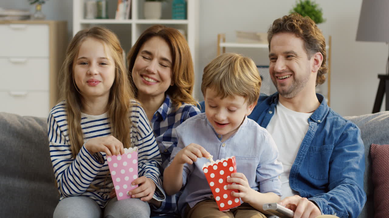 счастливая улыбающаяся семья с попкорном сидит на диване в гостиной и смеется, смотря комедийный фильм