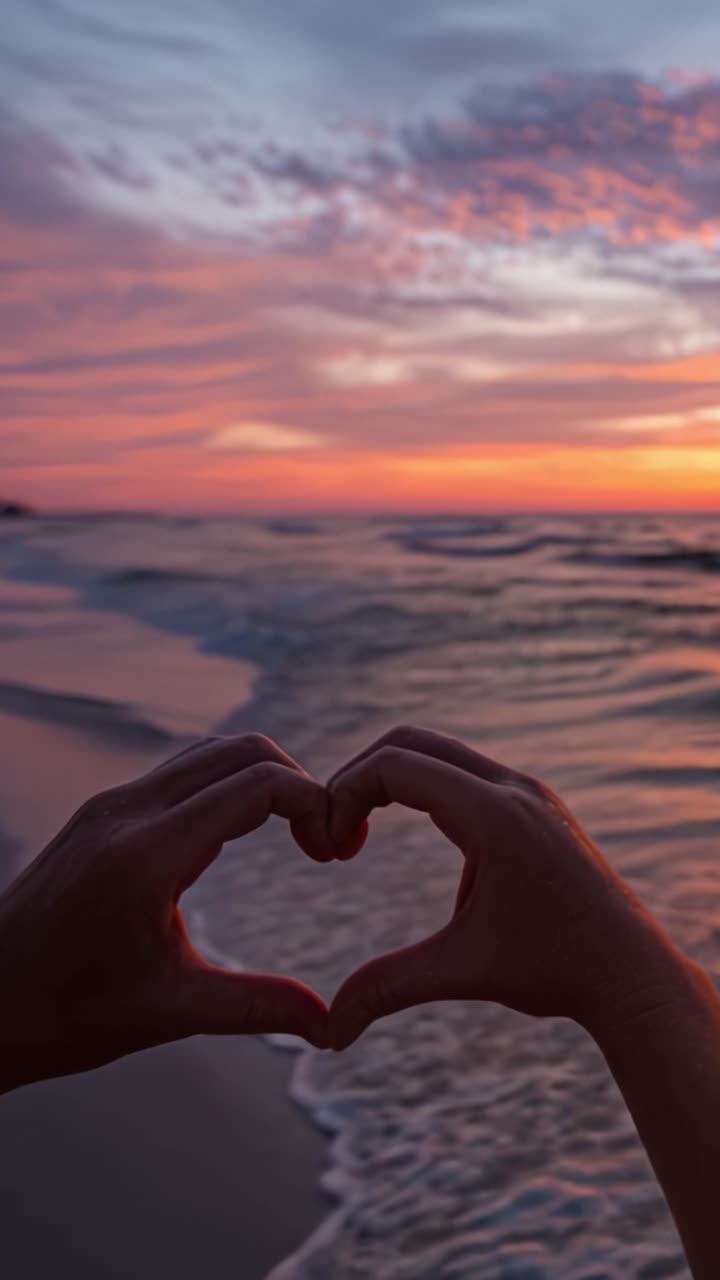Uno splendido tramonto sull'oceano con l'amore simboleggiato da mani che formano un cuore, mentre le onde si infrangono dolcemente sulla riva, catturando un momento di serenità e affetto.