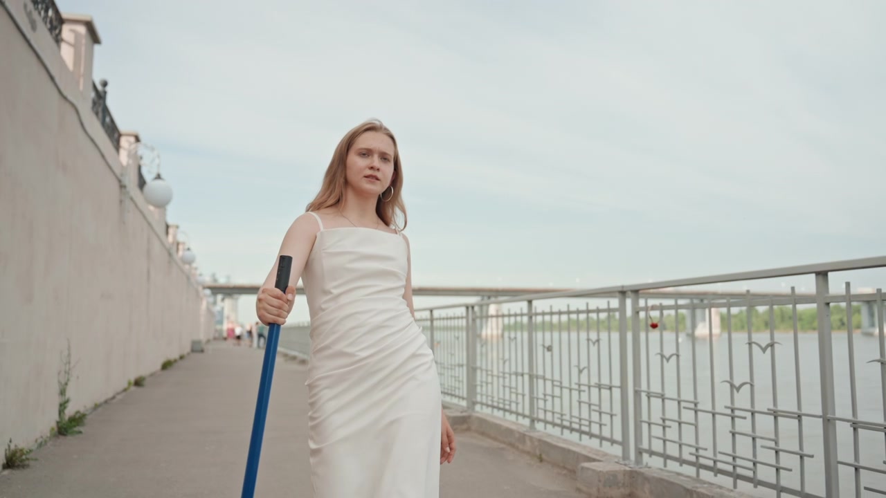 Una donna in abito bianco cammina lungo una passeggiata fluviale, tenendo un oggetto e guardandosi intorno. La scena presenta acqua calma, vegetazione distante, ringhiere metalliche con disegni di uccelli e una luce diurna soffusa.
