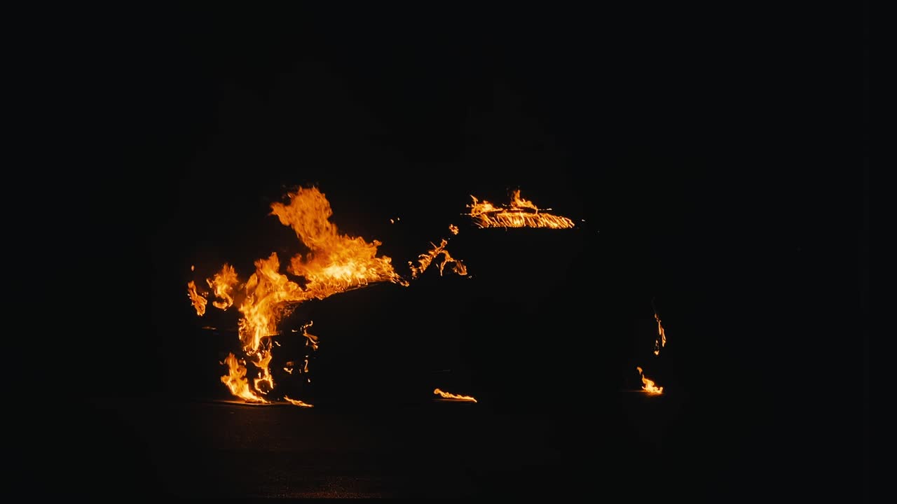 Car engulfed in flames at night with dark background burns until team of firefighters extinguish the flames with water hose.
