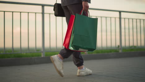 leg view of lady in grey joggers walking along iron rail with shopping bag in hand and black handbag, featuring soft blur background with distant greenery