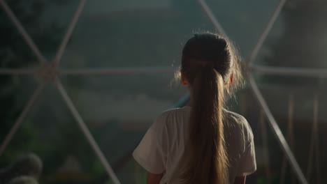 child walks under glass dome at glamping. little girl observes transparent wall with metal frame in hotel. kid looks at reflection in window