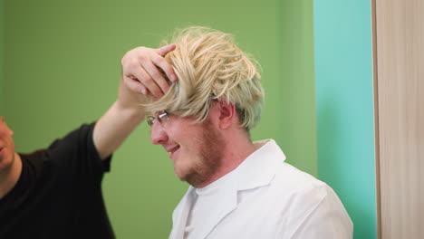 a close view of a man in a white lab coat wearing a blonde wig and black gloves, while another person in a black polo shirt playfully adjusts the wig, with bright green background