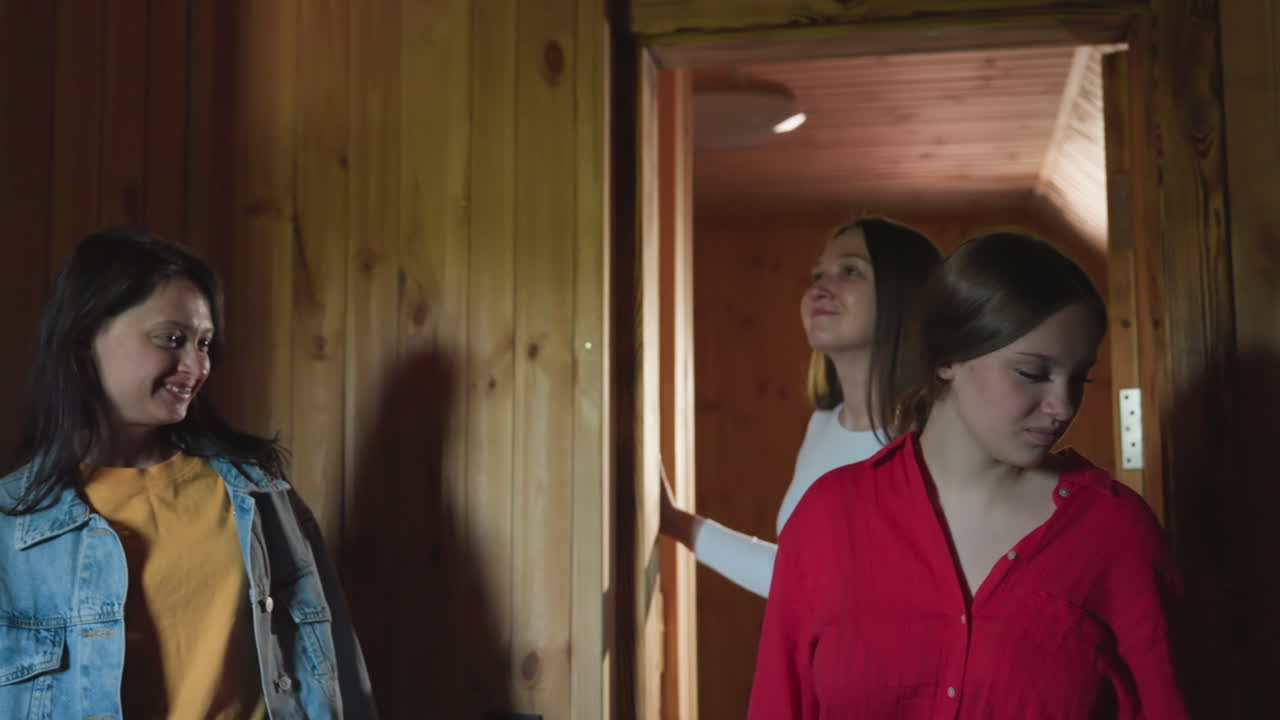 woman in red shirt enters rustic wooden lounge with friend, pausing by door to survey cozy interior in daylight, conveying exploration curiosity during first visit to charming cabin retreat