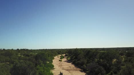 Toma-De-Un-Dron-En-Aumento-De-Vehículos-De-Juego-En-Un-Lecho-De-Río-Seco-En-El-Calor-Del-Sol-De-La-Tarde-Africana-Rastreando-Animales