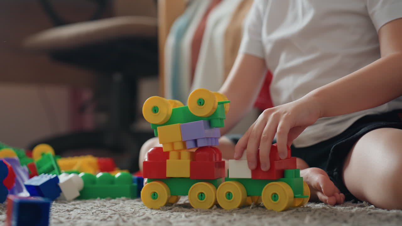 Un bambino bianco costruisce un treno di blocchi colorati. Primo piano di mani che assemblano mattoncini di plastica su un tappeto, ruote giallo brillante, impilamento attento. Gioco calmo e concentrato in una camera da letto accogliente con luce naturale soffusa.